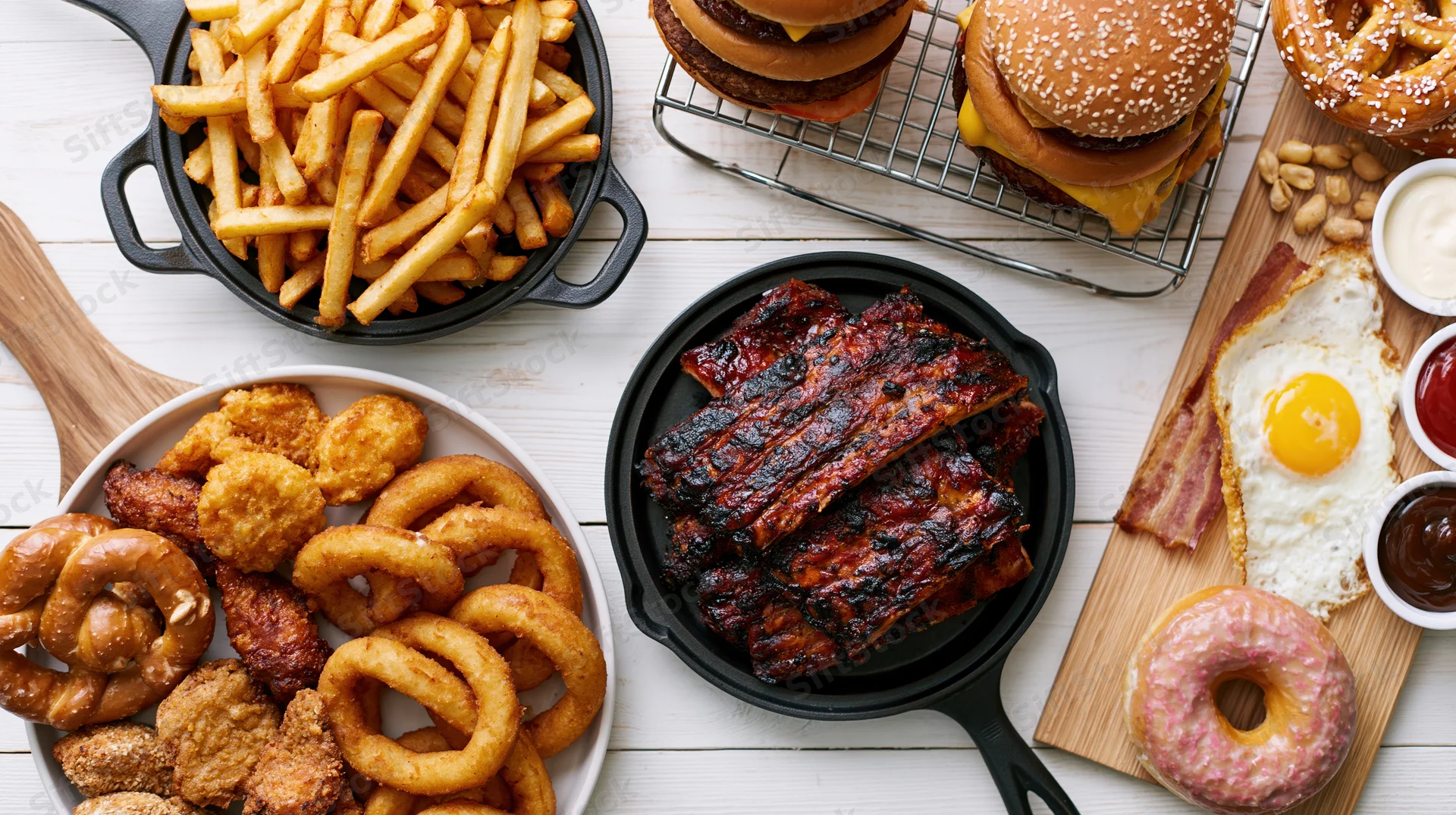 Overhead flat lay of american comfort food spread on white wooden table featuring burgers, fries, ribs, onion rings, pretzels, donut, fried egg, bacon, and condiments