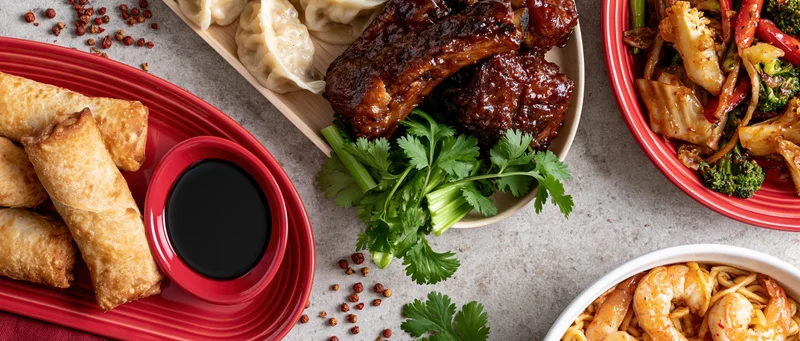 Overhead flat lay of an asian food spread with spring rolls, ribs, dumplings, stir fry, and noodles on a gray stone surface