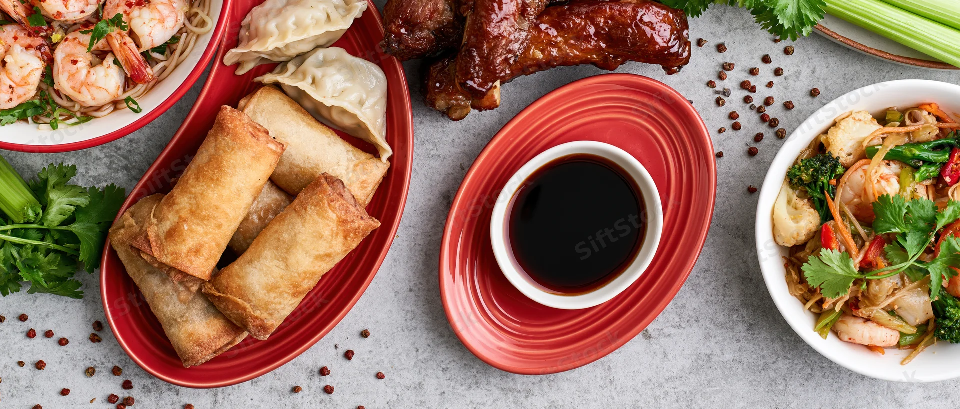 Overhead flat lay of an asian food spread with spring rolls, dumplings, shrimp noodles, stir fry, and soy sauce on a gray surface