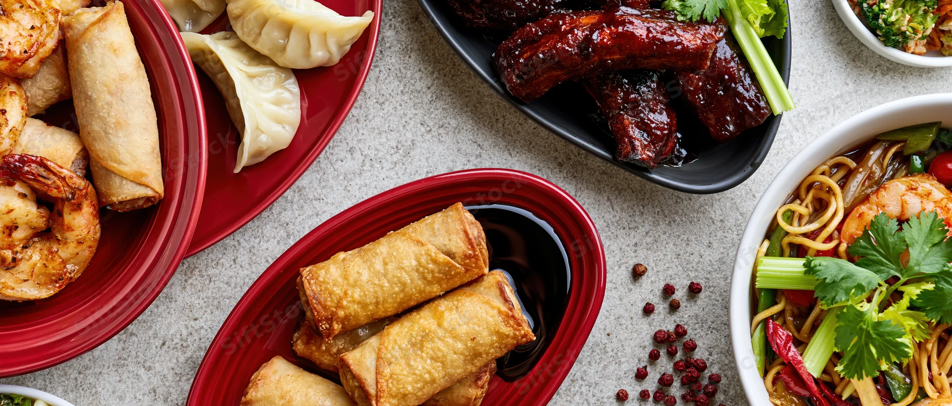 Overhead flat lay of an asian food spread with spring rolls, dumplings, ribs, noodles, and shrimp on red and black plates on a light gray surface