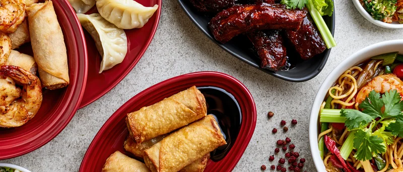 Overhead flat lay of an asian food spread with spring rolls, dumplings, ribs, noodles, and shrimp on red and black plates on a light gray surface