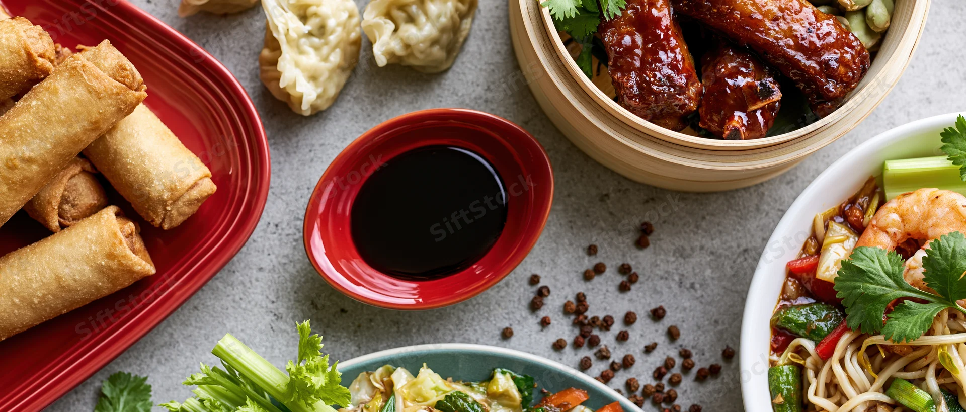 Overhead flat lay of an asian food spread with spring rolls, dumplings, glazed wings, noodles, and soy sauce on a grey surface