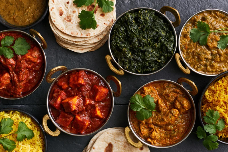 Overhead flat lay of an indian feast spread across a dark slate table with multiple curries, rice, naan, and garnishes in metal bowls