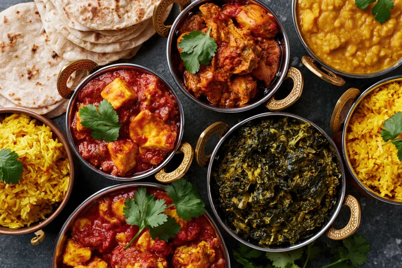 Overhead flat lay of an indian feast with curry, rice, naan and vegetables served in brass bowls on a dark surface