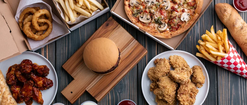 Overhead flat lay of assorted american fast food spread across wooden table with burger, pizza, fries, fried chicken, onion rings, and sauces on plates and boxes