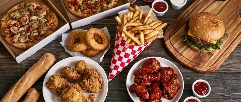 Overhead flat lay of assorted american fast food spread across wooden table with burger, pizza, fries, chicken wings, onion rings, and condiments