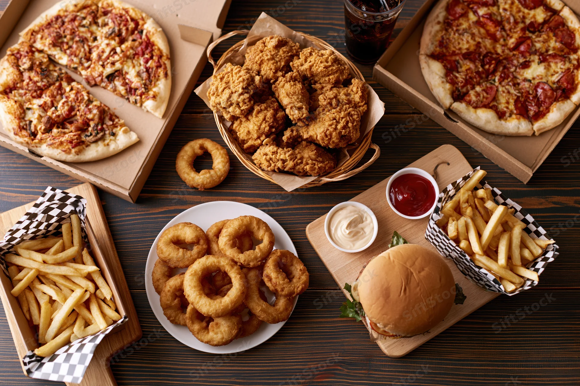 Overhead flat lay of assorted american fast food spread across dark wood table including pizza, burger, fries, onion rings, fried chicken, and soda