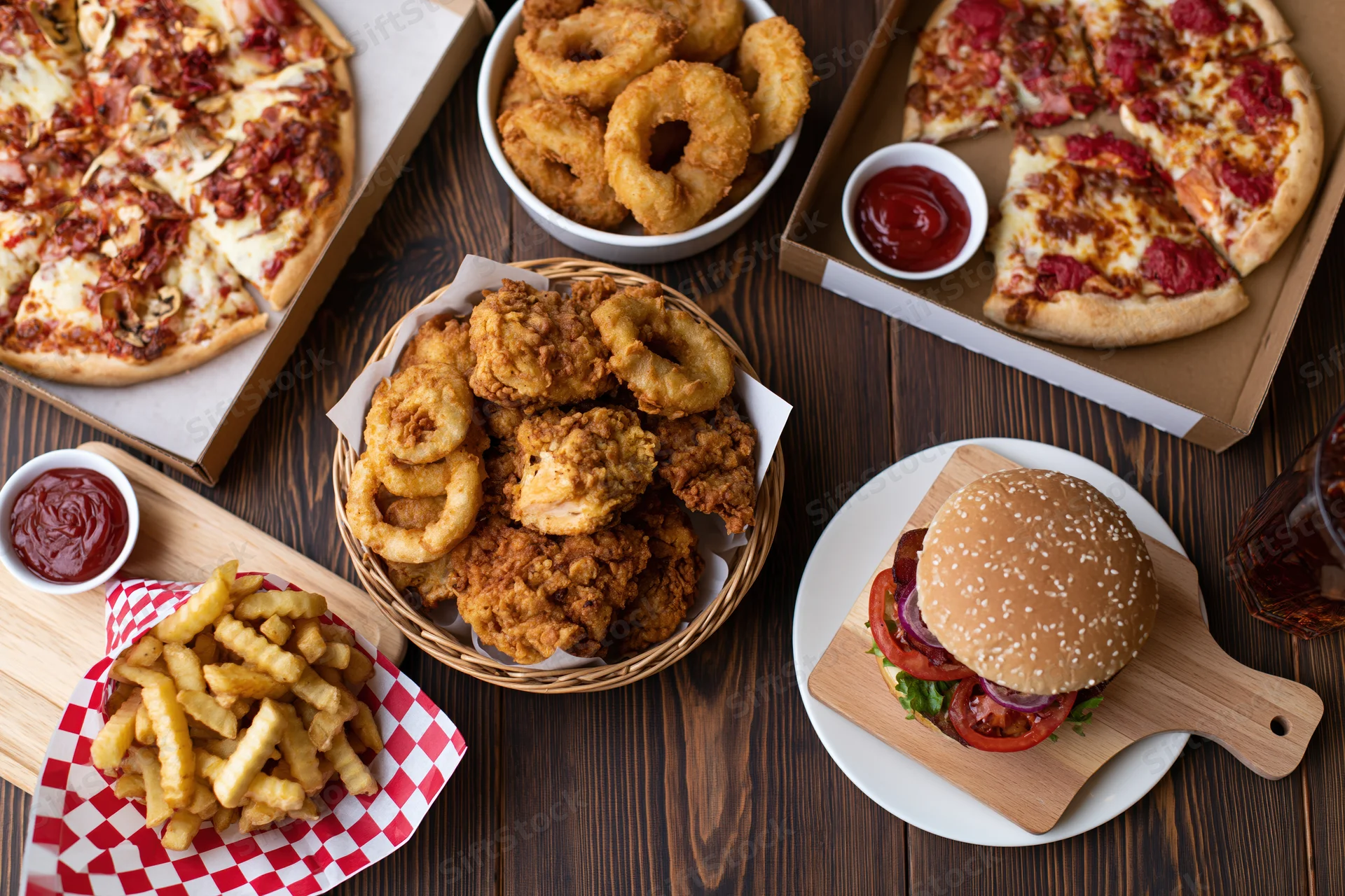 Overhead flat lay of assorted american fast food including pizza, burger, fries, onion rings and chicken on dark wood surface