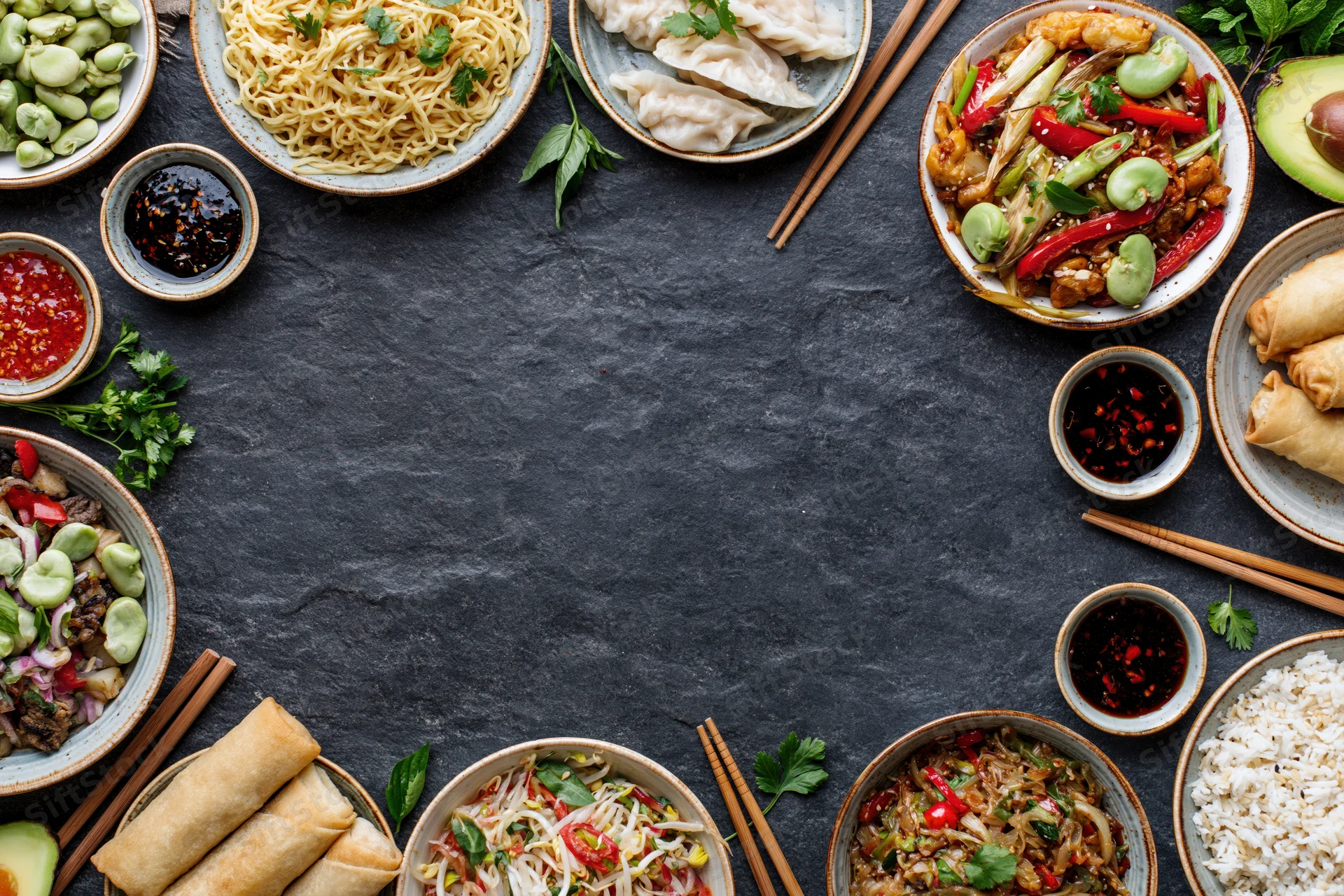Assorted asian dishes arranged in a circular flat lay on dark slate surface with copyspace in center