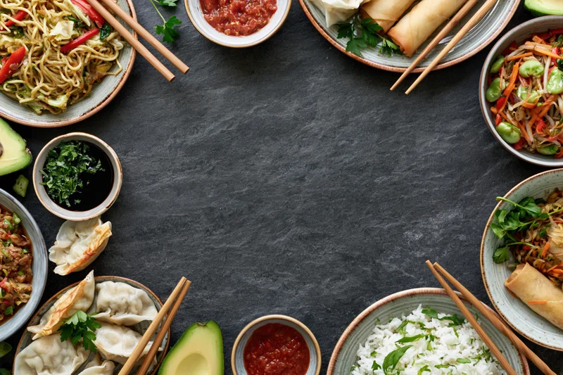 Assorted asian dishes arranged in a circular flat lay on dark slate surface with chopsticks and garnishes