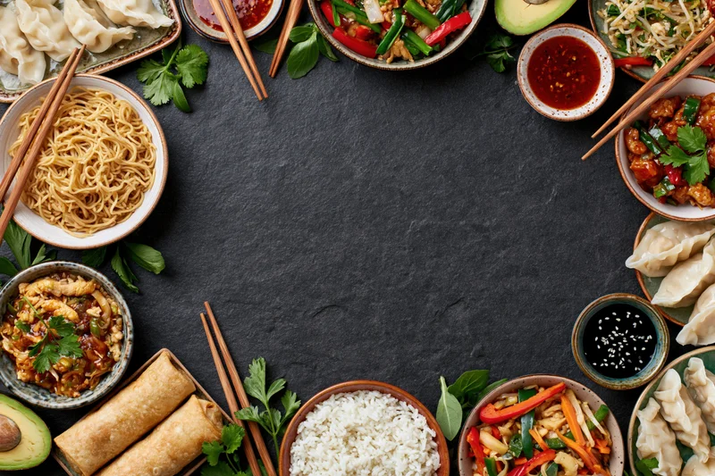Assorted asian dishes arranged in a circular flat lay on a dark slate surface with chopsticks and garnishes, leaving central copyspace