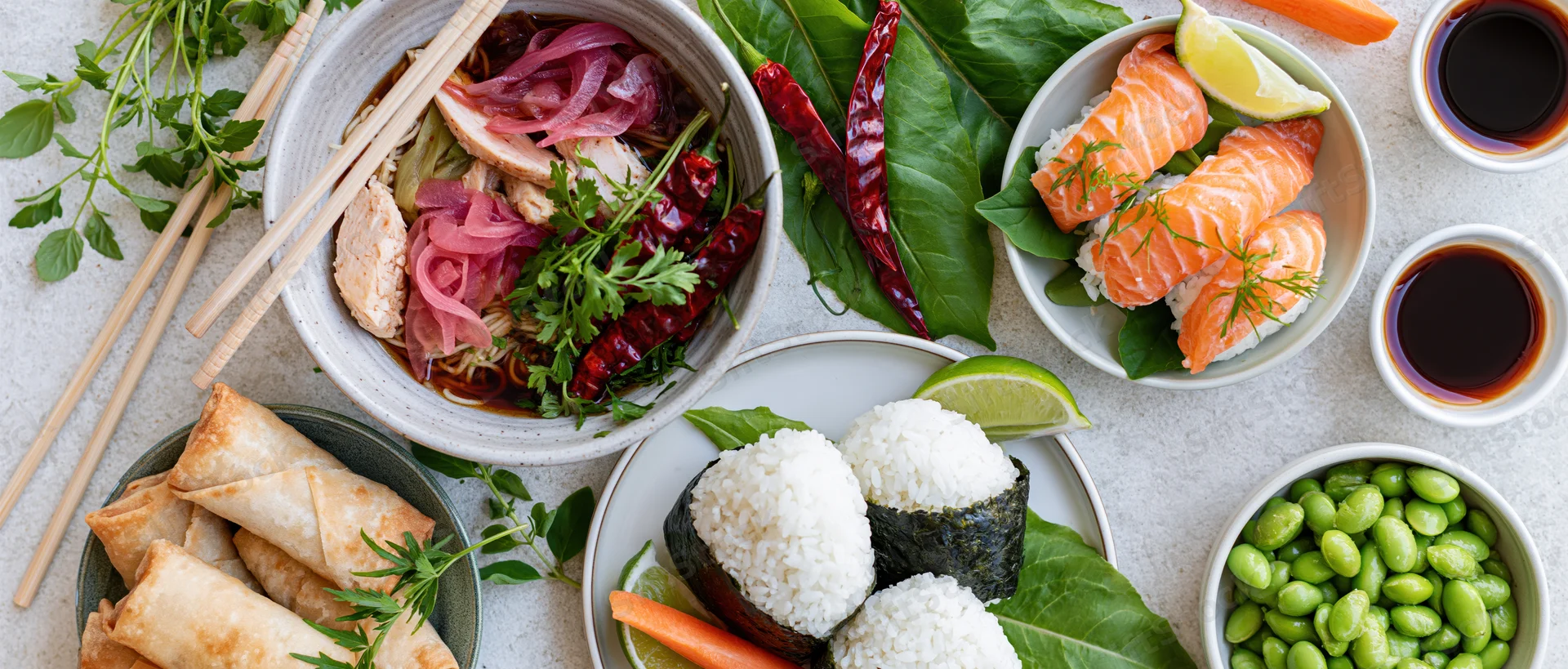 Overhead flat lay of assorted Asian dishes including sushi, ramen, spring rolls, edamame, and soy sauce on a light concrete surface