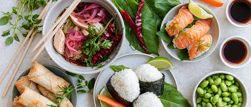 Overhead flat lay of assorted Asian dishes including sushi, ramen, spring rolls, edamame, and soy sauce on a light concrete surface