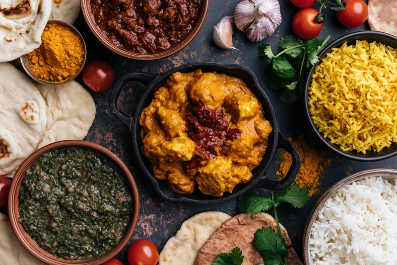 Overhead flat lay of assorted indian dishes including curry, rice, naan, and spices arranged on a dark textured surface
