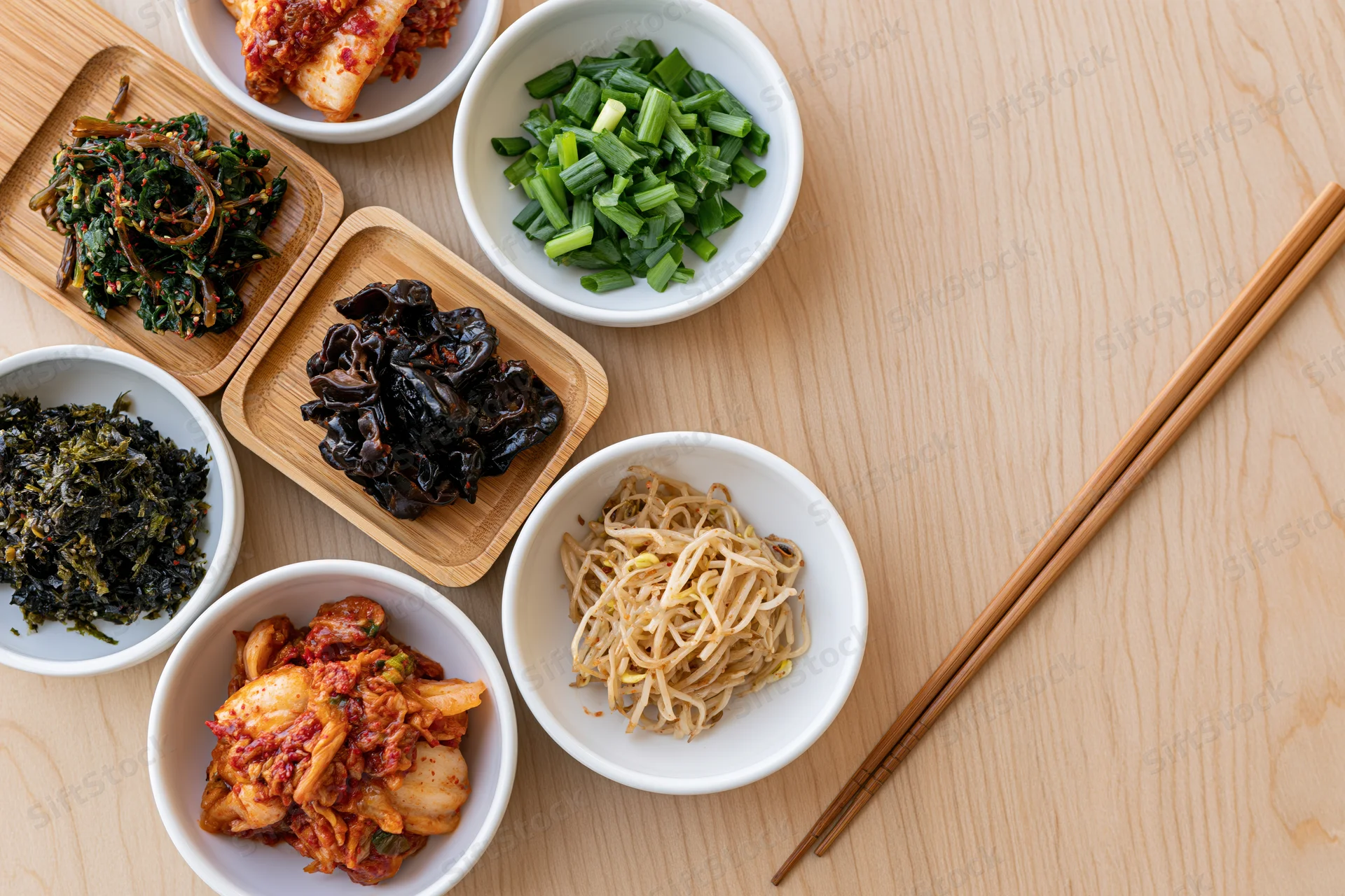 Overhead flat lay of assorted korean banchan side dishes in white bowls and wooden trays with chopsticks on a light wood table
