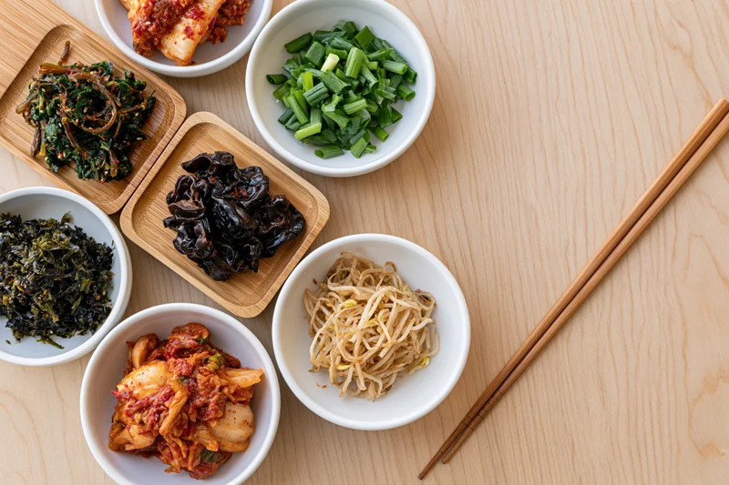 Overhead flat lay of assorted korean banchan side dishes in white bowls and wooden trays with chopsticks on a light wood table