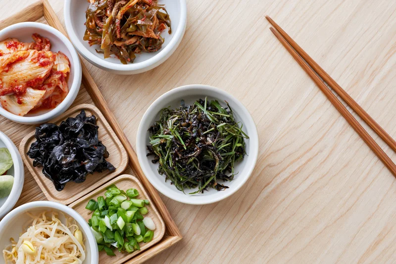Assorted korean banchan side dishes arranged on a wooden tray with chopsticks on a light wood table