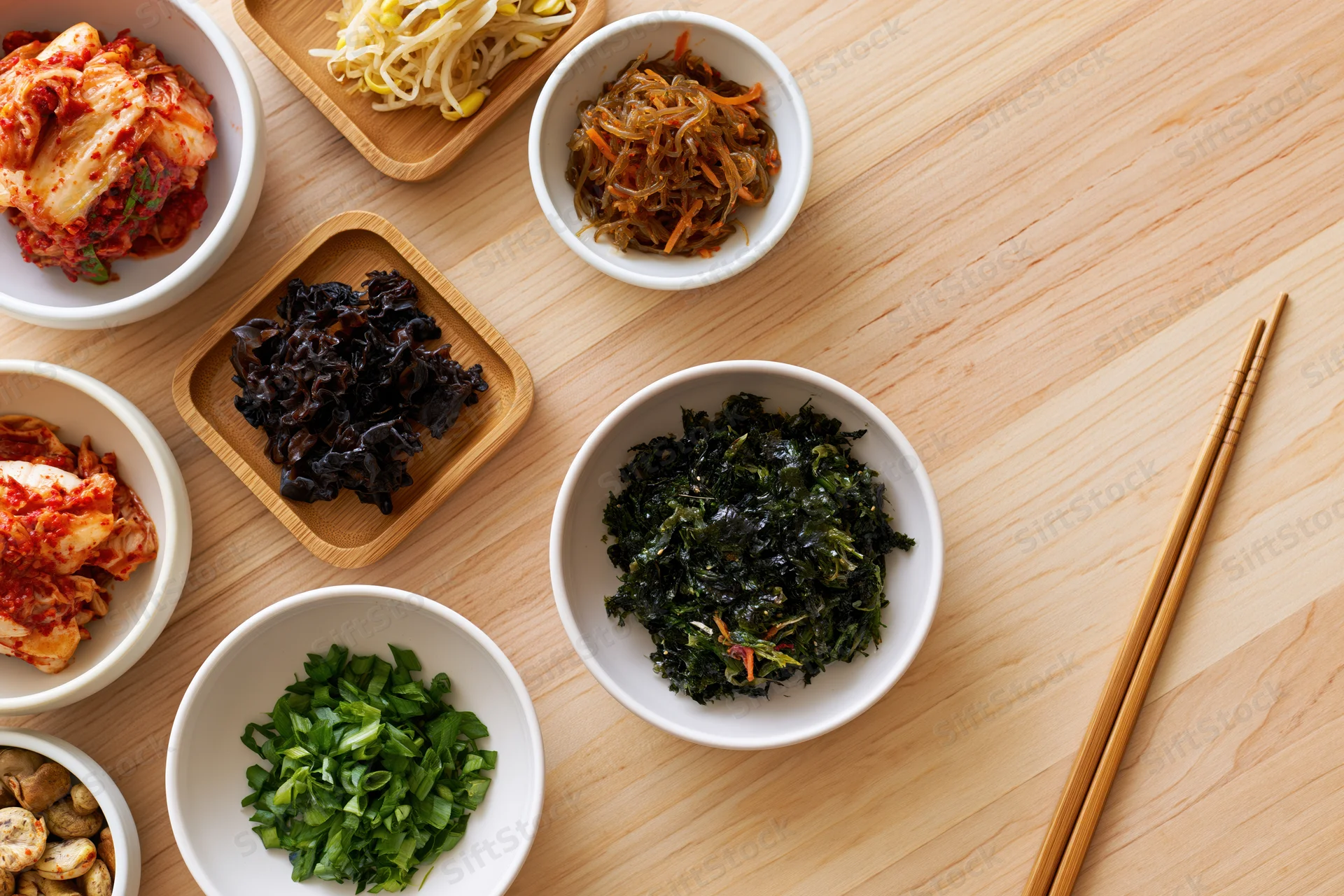 Overhead flat lay of assorted korean banchan side dishes arranged on wooden table with chopsticks and white bowls