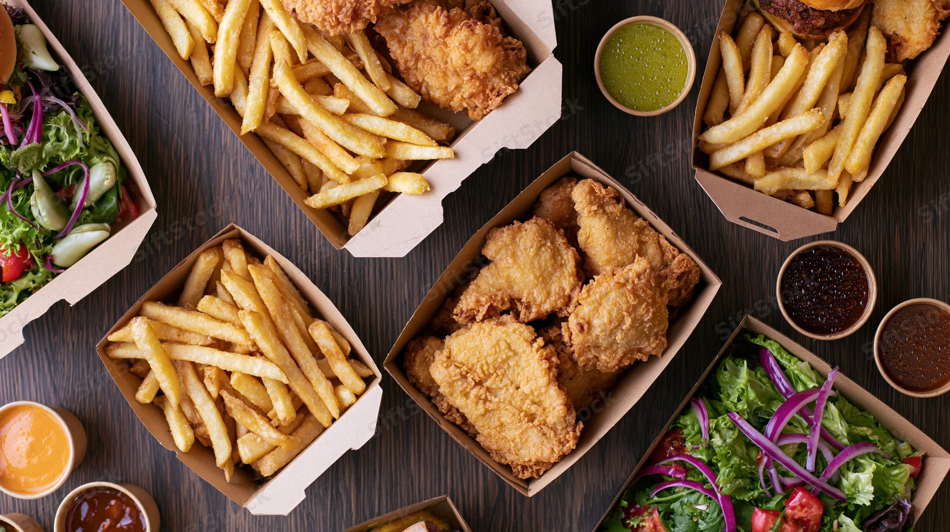 Overhead flat lay of assorted fast food including fried chicken, french fries, and salads arranged on dark wood surface with dipping sauces in small bowls