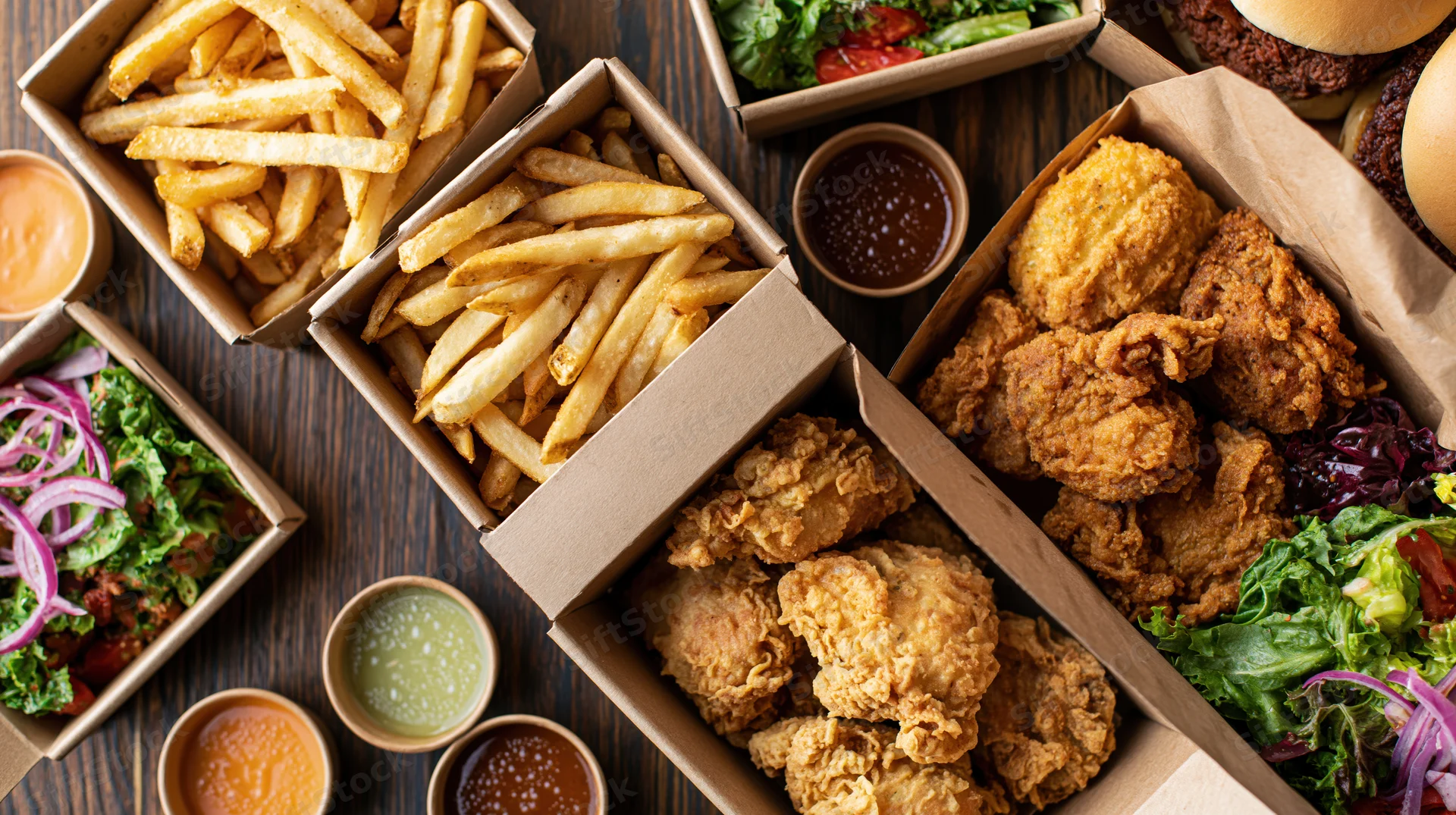 Fried chicken and french fries in cardboard boxes with salads and dipping sauces arranged on dark wood surface