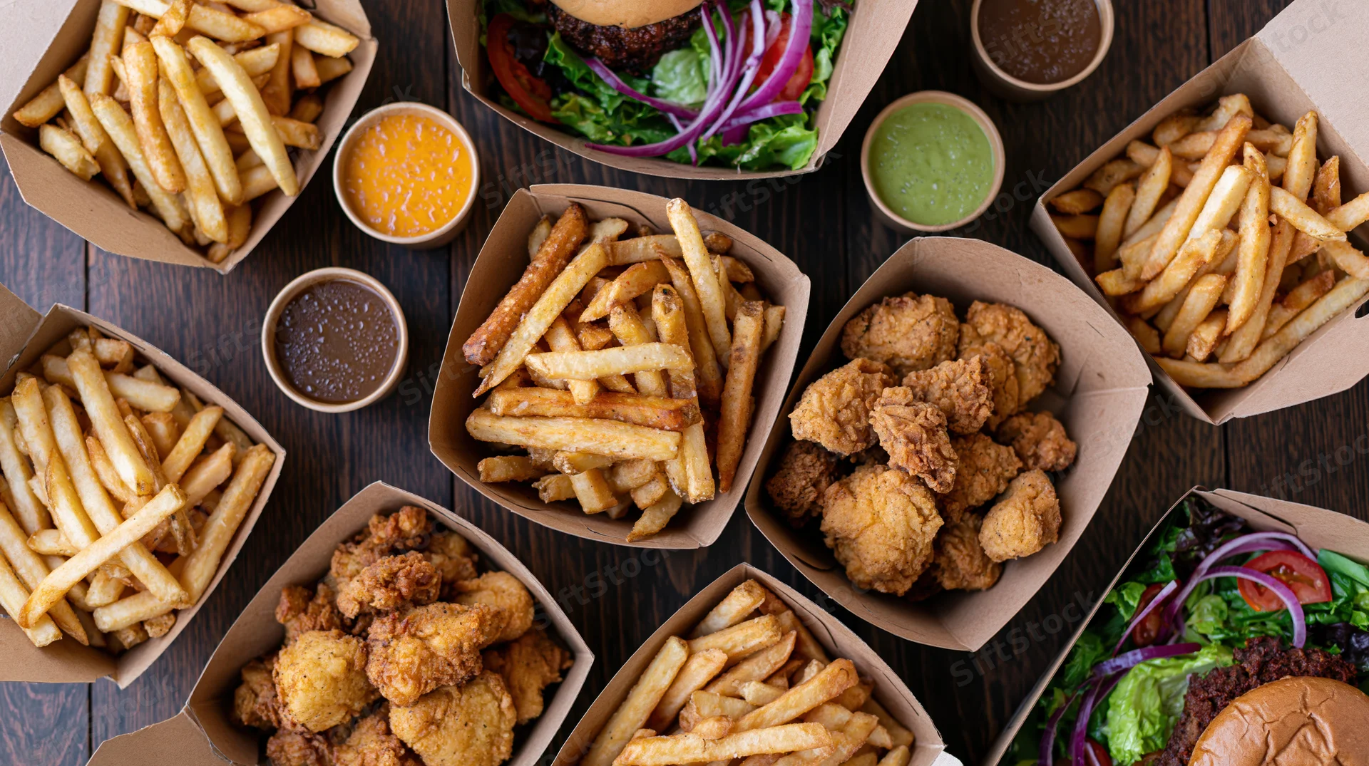 Overhead flat lay of assorted fast food including burgers, fries, and fried chicken on a dark wooden table