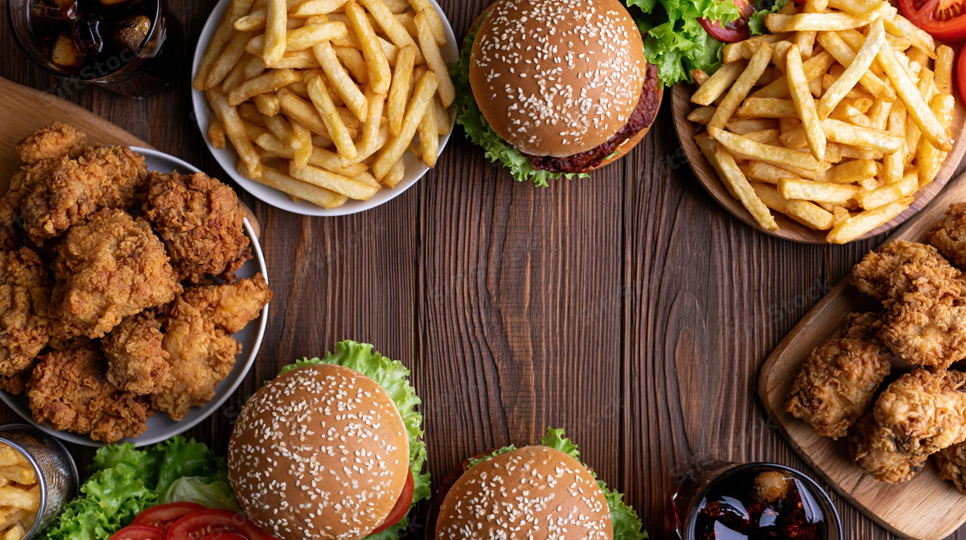 Overhead flat lay of burgers, fries, fried chicken and soda arranged on dark wooden table