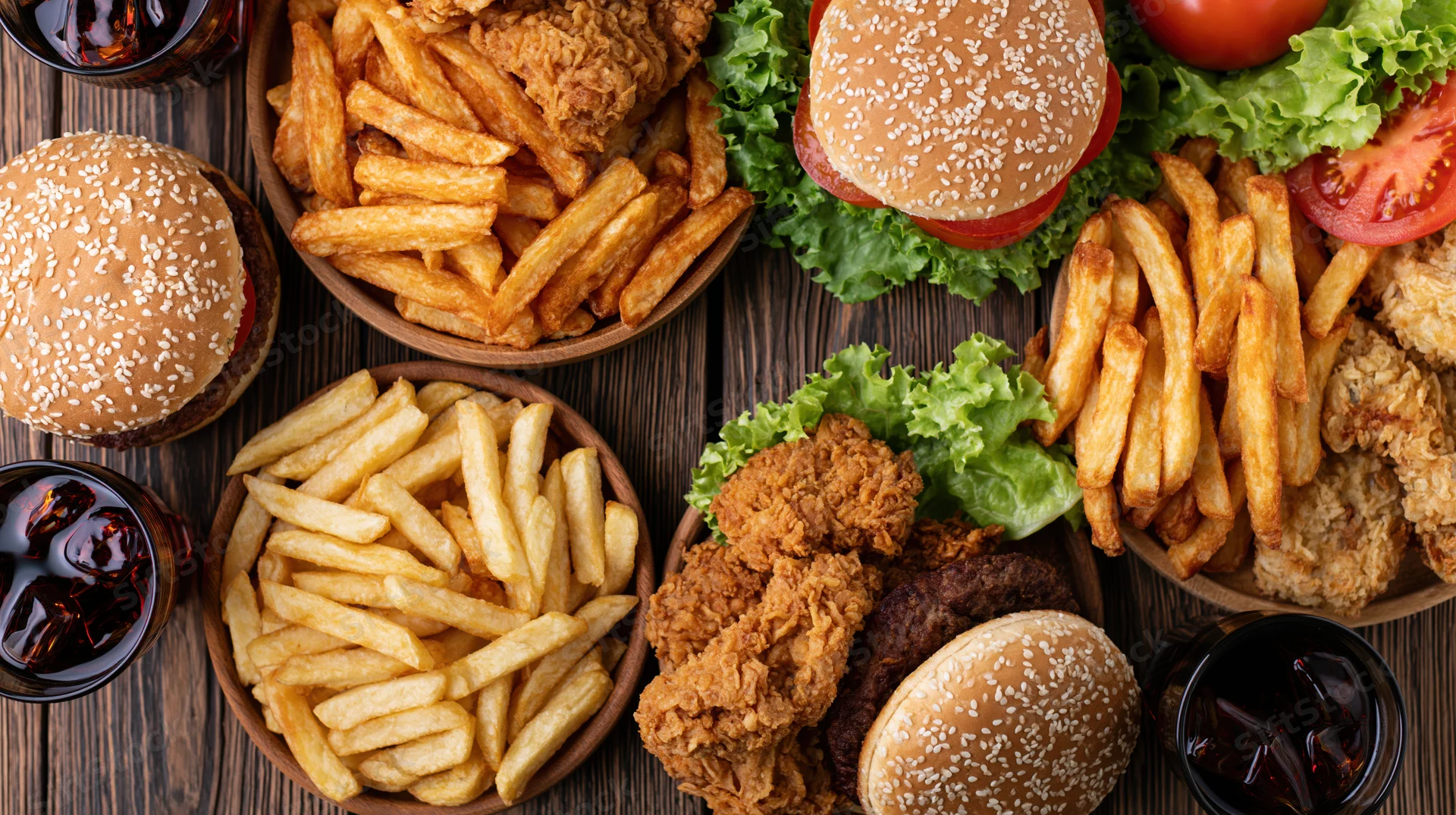 Overhead flat lay of assorted fast food including burgers, fries, fried chicken and soda on a dark wooden table
