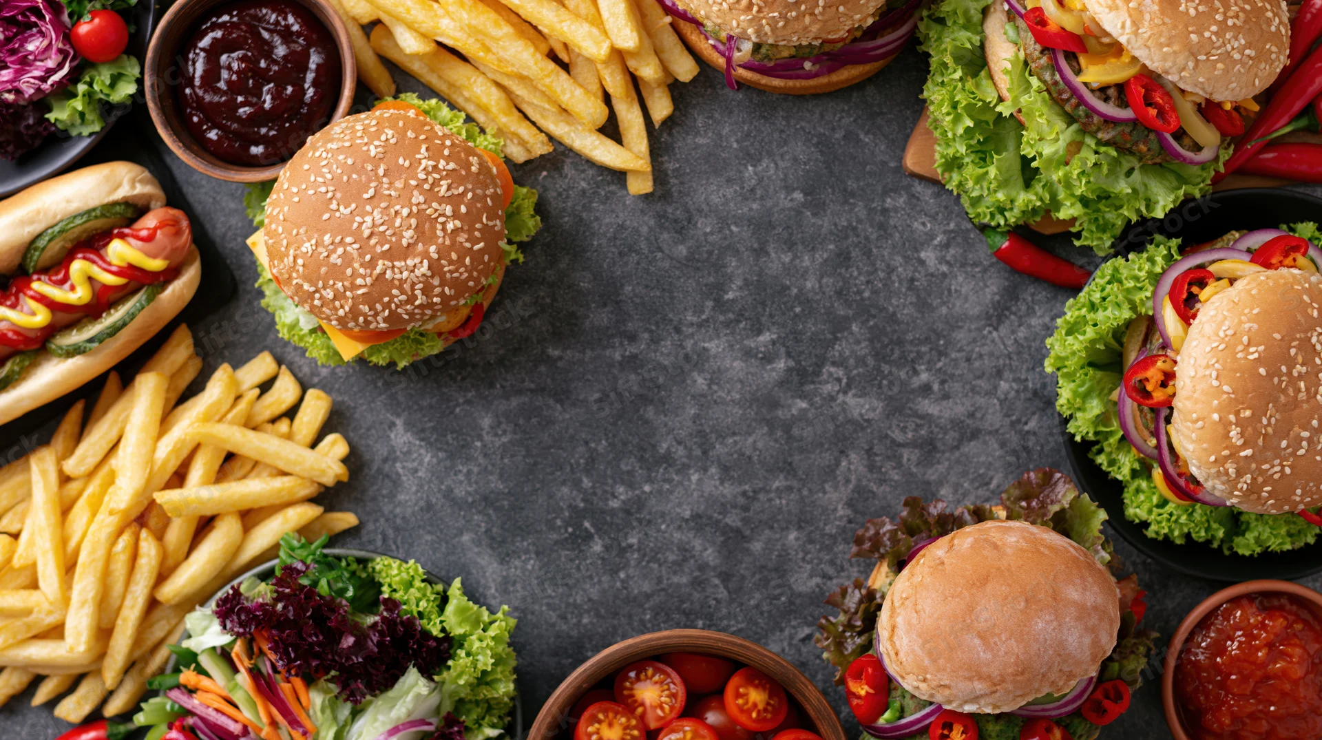 Assorted fast food items arranged in a circular flat lay on a dark stone surface with central copyspace
