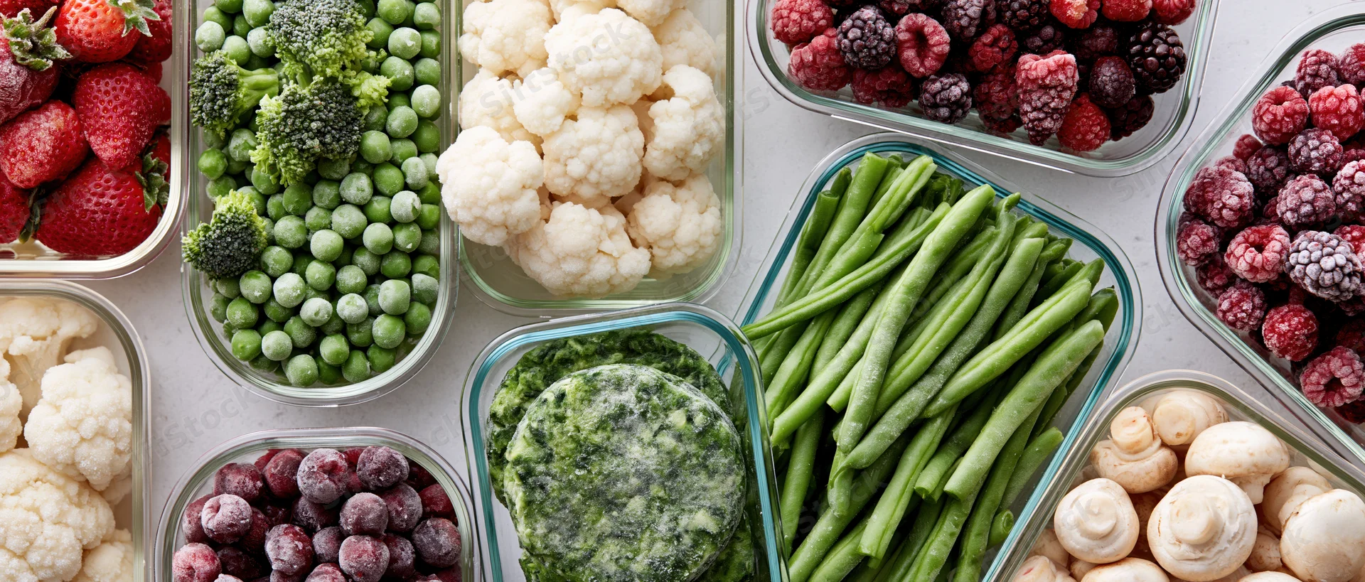 Overhead flat lay of assorted frozen vegetables and fruits in glass containers on a white countertop