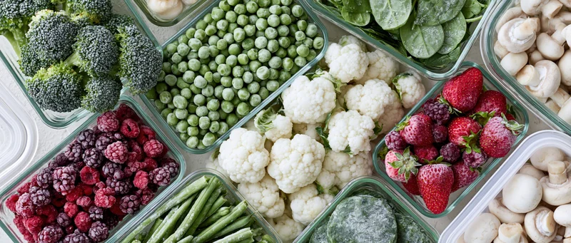 Overhead flat lay of assorted frozen vegetables and fruits in glass containers arranged in a grid pattern