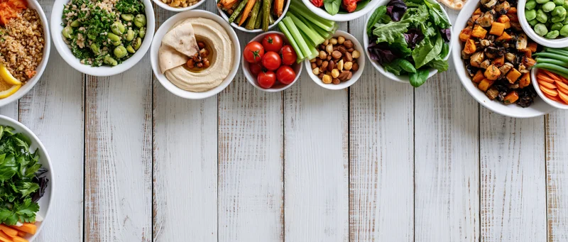 Overhead flat lay of assorted plant-based meals in white bowls on a wooden table
