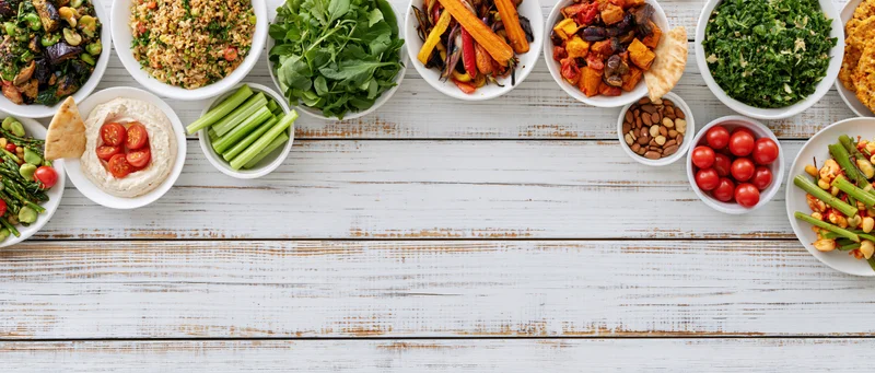 Overhead flat lay of assorted healthy plant-based meals and snacks arranged on a white wooden table with copyspace