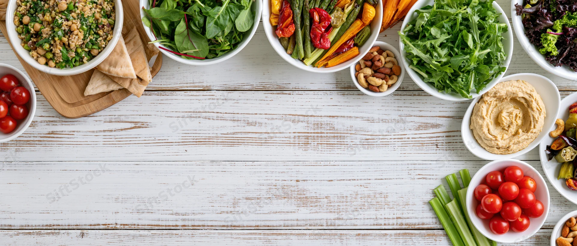 Overhead flat lay of assorted plant-based meals and snacks arranged on a white wooden table with copyspace