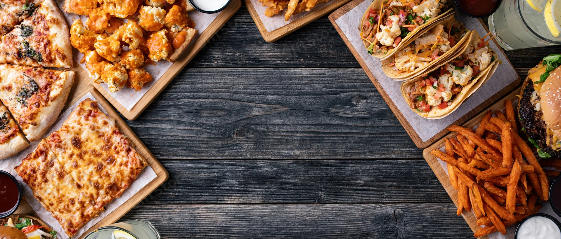 Assorted restaurant food arranged in a flat lay on a dark wooden table with copyspace in the center
