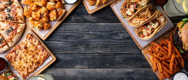 Assorted restaurant food arranged in a flat lay on a dark wooden table with copyspace in the center