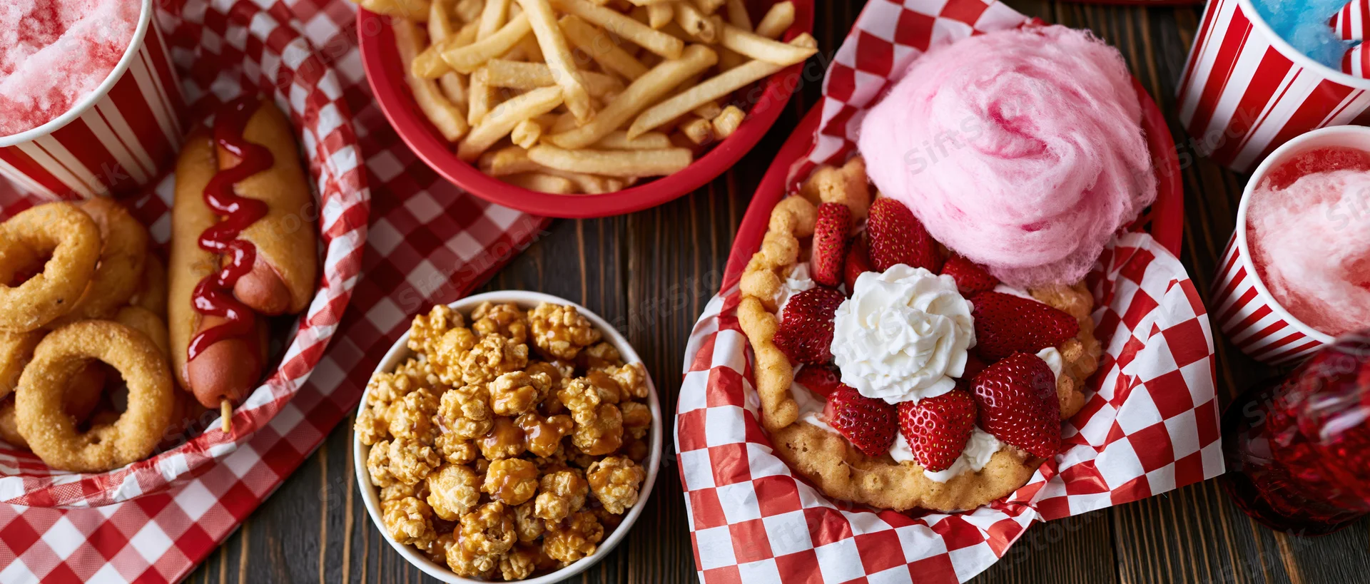 Overhead flat lay of classic american carnival fair food spread on wooden table with red and white checkered paper lining baskets and bowls