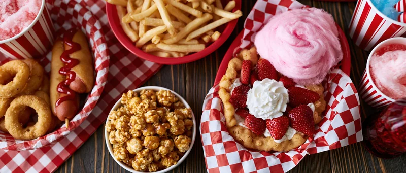 Overhead flat lay of classic american carnival fair food spread on wooden table with red and white checkered paper lining baskets and bowls