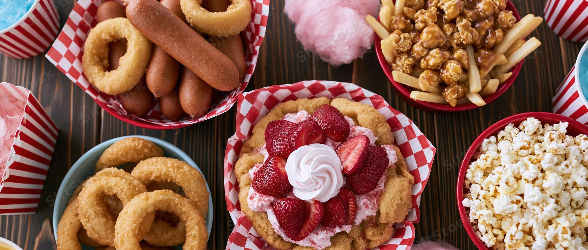 Overhead flat lay of classic american carnival fair food spread on wooden table with red and white checkered paper baskets