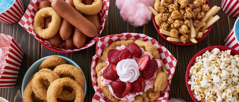 Overhead flat lay of classic american carnival fair food spread on wooden table with red and white checkered paper baskets