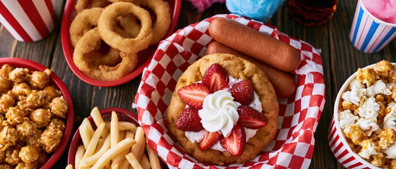Overhead flat lay of classic american carnival fair food spread on dark wooden table with red and white checkered paper baskets and striped cups