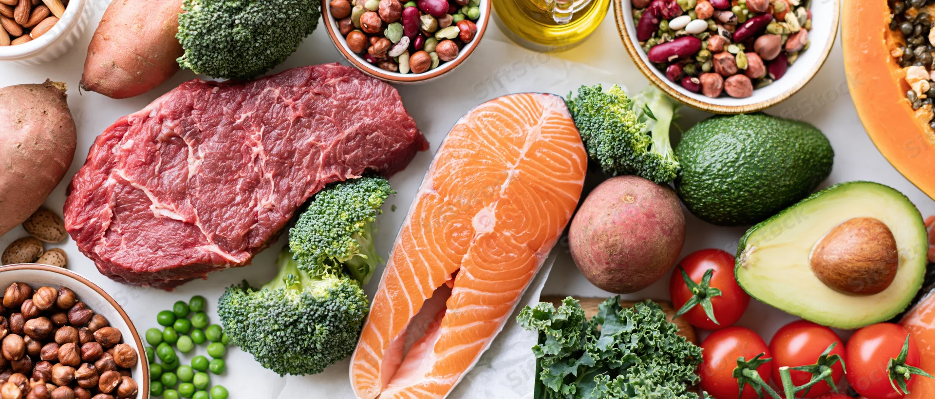Overhead flat lay of whole foods including salmon, beef, broccoli, avocado, sweet potato, tomato, kale, peas, hazelnuts, beans, pumpkin, and olive oil arranged on a white surface