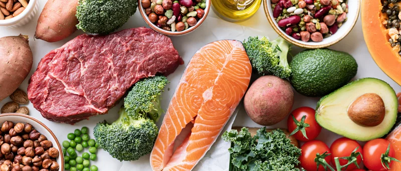 Overhead flat lay of whole foods including salmon, beef, broccoli, avocado, sweet potato, tomato, kale, peas, hazelnuts, beans, pumpkin, and olive oil arranged on a white surface