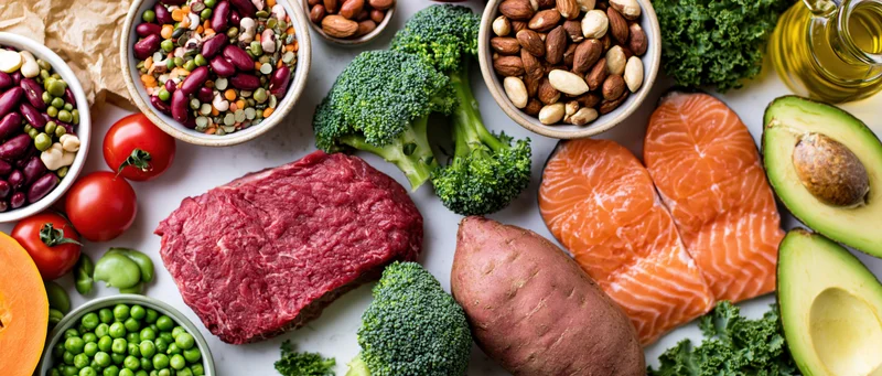 Overhead flat lay of diverse whole foods including salmon, beef, avocado, broccoli, sweet potato, tomatoes, beans, almonds, and olive oil arranged on a white surface