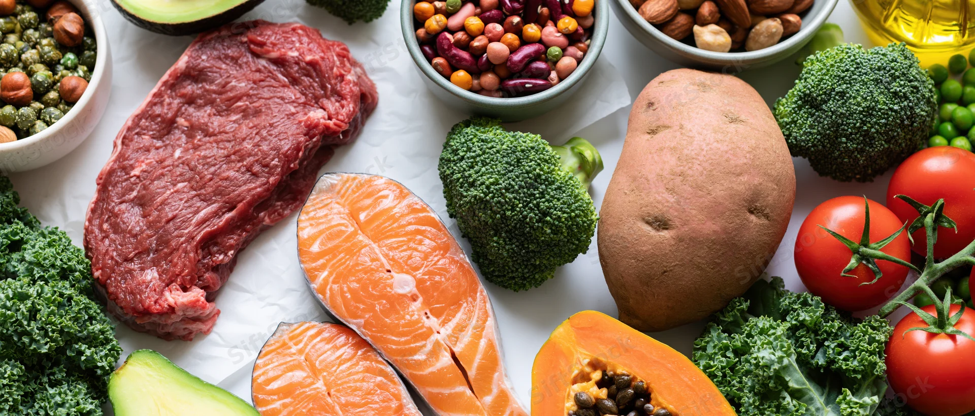 Overhead flat lay of diverse whole foods including salmon, beef, broccoli, sweet potato, tomato, avocado, kale, papaya, beans, nuts, and olive oil arranged on white surface