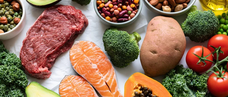 Overhead flat lay of diverse whole foods including salmon, beef, broccoli, sweet potato, tomato, avocado, kale, papaya, beans, nuts, and olive oil arranged on white surface
