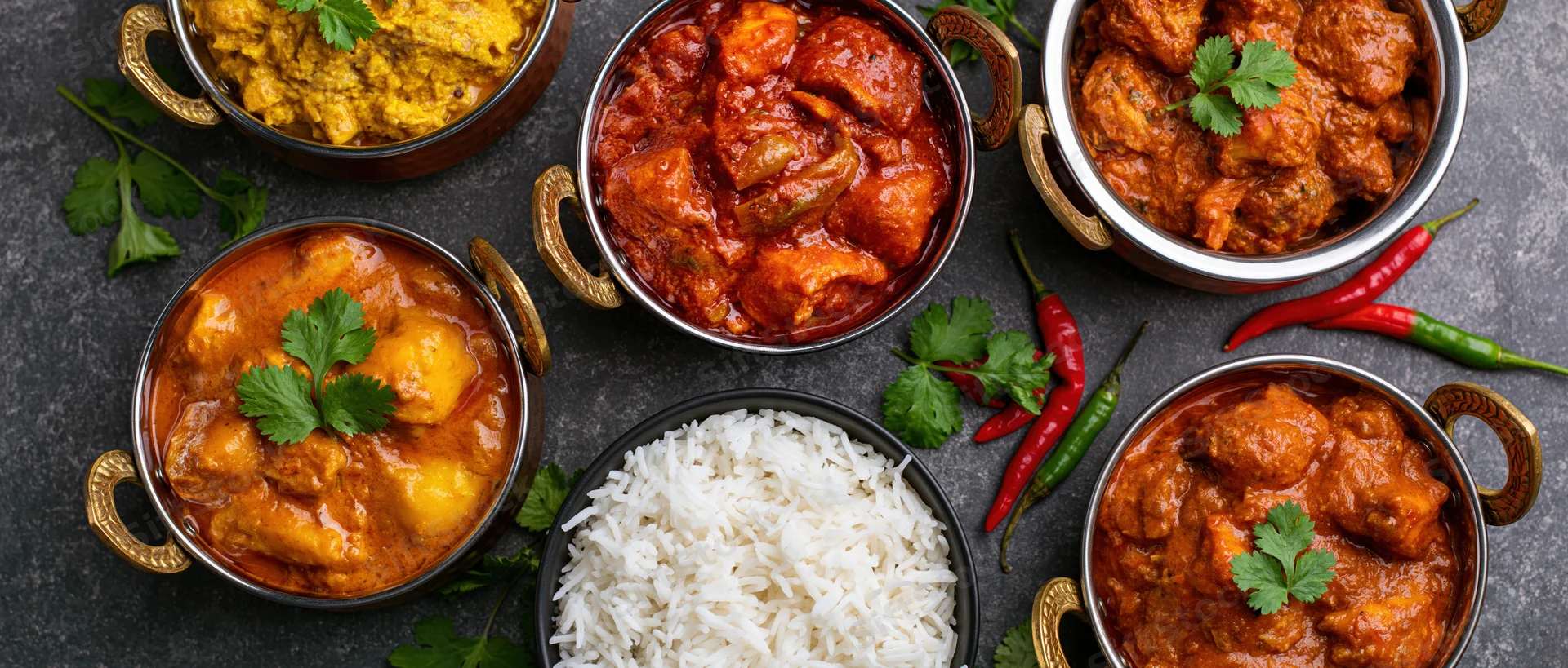 Overhead flat lay of multiple indian curry dishes and rice arranged on dark surface