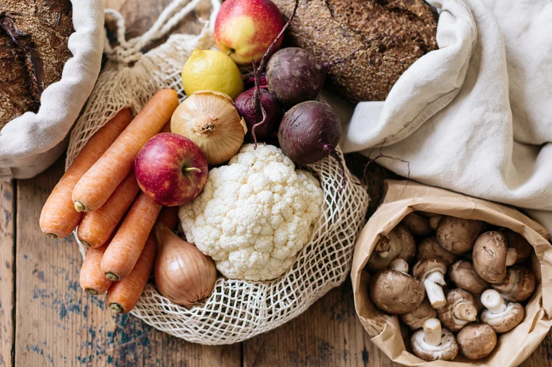 Fresh vegetables and bread arranged in reusable bags on a wooden surface
