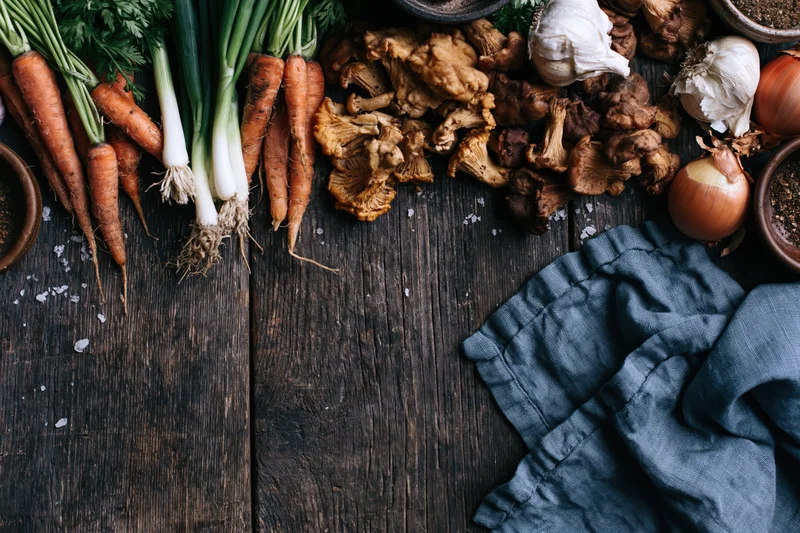 Fresh vegetables and mushrooms arranged on a dark wooden table with a blue cloth and salt sprinkled around