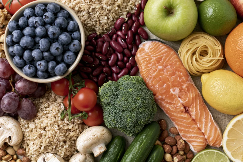 Overhead flat lay of fresh whole foods densely arranged on a surface including salmon, broccoli, blueberries, brown rice, kidney beans, apple, lemon, orange, lime, grapes, tomatoes, mushrooms, cucumbers, nuts, and pasta
