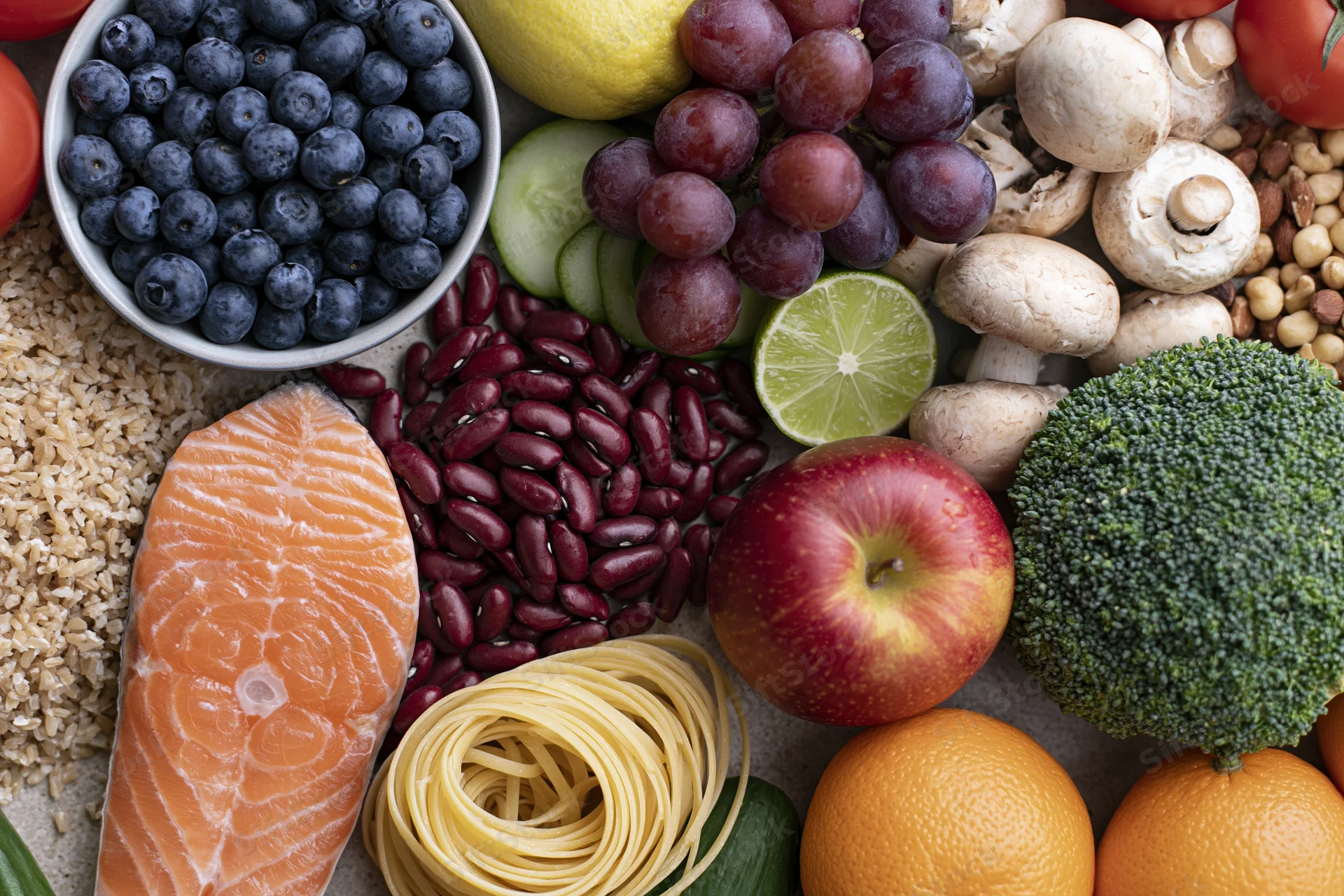 Overhead flat lay of fresh whole foods densely arranged on a surface including salmon, rice, beans, pasta, broccoli, apple, orange, lime, grape, blueberry, tomato, cucumber, mushroom, nuts, and seeds